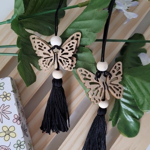 Peut inclure: Deux breloques en bois en forme de papillon avec des glands noirs suspendus à une surface en bois. Les papillons sont brun clair et présentent des découpes complexes. Les glands sont en ficelle noire et mesurent environ 10 cm de long.