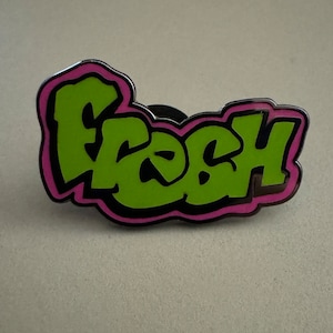 May include: A green and pink enamel pin with the word "Fresh" in a graffiti style font.