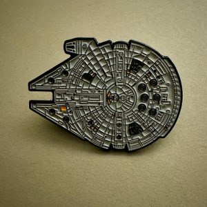 May include: A black and white metal pin of the Millennium Falcon spaceship from Star Wars. The pin is detailed with the ship's cockpit and engine details.