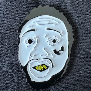 May include: A black and white enamel pin featuring a cartoon face with a gold grill and a black and white tattoo on the cheek.