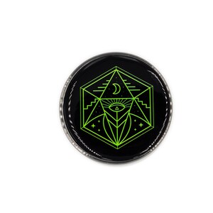 Op de afbeelding: Een ronde, zwarte knoop met een geometrisch ontwerp in neon groen. Het ontwerp bevat een maan, een oog en stervormige elementen. De knoop heeft een zilverkleurige metalen rand en staat tegen een witte achtergrond.