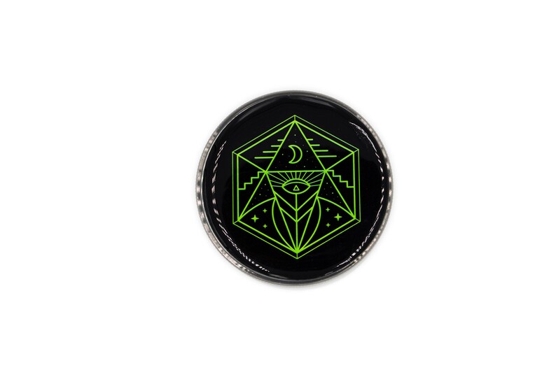 Op de afbeelding: Een ronde, zwarte knoop met een geometrisch ontwerp in neon groen. Het ontwerp bevat een maan, een oog en stervormige elementen. De knoop heeft een zilverkleurige metalen rand en staat tegen een witte achtergrond.