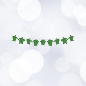 May include: A garland of green glitter turtle shapes suspended on a thin white string. The turtles are uniformly spaced and feature a simple, stylised design. The background is a soft, out-of-focus pattern of white and light blue.