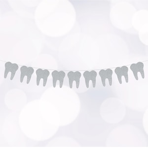 May include: A silver glitter banner with tooth-shaped cutouts strung on a thin white string. The teeth are evenly spaced and have a sparkly texture. The background is a soft, out-of-focus white with a bokeh effect.