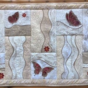 Op de afbeelding: Een beige en witte gequilte wandhanger met een patchwork-ontwerp met geborduurde vlinders en bloemen in tinten rood. Het ontwerp is abstract en heeft vloeiende lijnen en swirls.