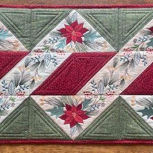 May include: A rectangular quilted table runner with a Christmas theme. The design features red poinsettias, green triangles, and diagonal red stripes. The runner is bordered with a red trim and has a festive, holiday aesthetic.