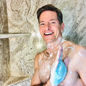 May include: A man in a shower with a blue washcloth in his hand. He is covered in soap suds and smiling at the camera.
