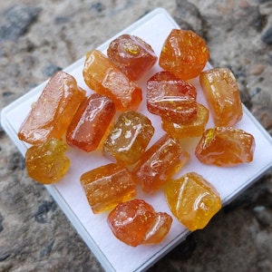 May include: A collection of small, translucent, amber-colored gemstones with irregular, faceted shapes. These stones exhibit varying shades of orange and yellow, featuring a glossy, polished surface texture that reflects light across their uneven edges.