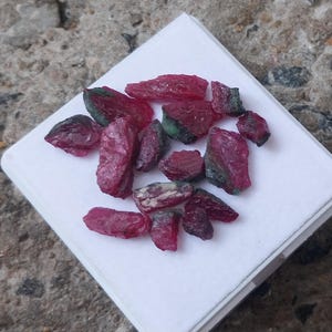 May include: A collection of raw, unpolished ruby gemstones with deep red and magenta hues, featuring irregular, jagged shapes and natural crystalline textures. These small, opaque mineral fragments are displayed together on a square white surface.