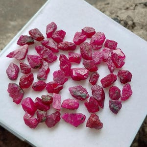 May include: A collection of raw, uncut ruby gemstones. The rubies vary in size and shape, displaying a deep red hue with some variations in color and texture. The gemstones are scattered on a white surface.