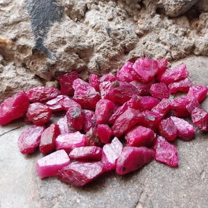 May include: A collection of raw, unpolished ruby gemstones in a vibrant, deep pinkish-red hue. These small, irregular mineral fragments feature a translucent to opaque appearance with natural facets and jagged edges, scattered across a surface.