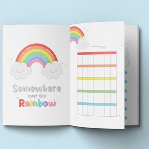 Puede incluir: Un cuaderno abierto con un arcoíris y nubes en la portada. El texto "Somewhere over the Rainbow" está escrito debajo del arcoíris. La página derecha tiene una cuadrícula de calendario en blanco con la palabra "mes" en la parte superior.