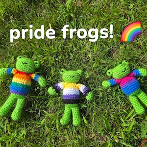 May include: Three handmade crochet frogs with green bodies and colorful striped tops, lying on green grass. The frogs' tops feature rainbow, non-binary, and bisexual pride flag colors. The image includes the text "pride frogs!" and a rainbow.