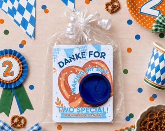 Peut inclure: Un sachet cadeau en plastique transparent contenant une carte rectangulaire avec une illustration de bretzel et le texte DANKE FOR MAKING MY DAY TWO SPECIAL! YOUR FRIEND, DAMIAN. Un couvercle circulaire en plastique bleu foncé est fixé.