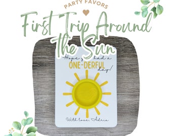 May include: A yellow sun-shaped party favor with the text "First Trip Around The Sun" and "Hope you had a ONE-DERFUL day!" with the signature "With love, Adria".