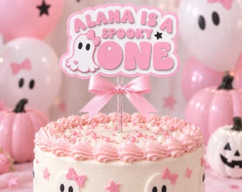 Peut inclure: Une décoration de gâteau avec le texte ALANA IS A SPOOKY ONE en lettres roses et une icône de fantôme blanc portant un nœud rose. La décoration est fixée sur un bâton transparent orné d'un nœud en ruban de satin rose, posée sur un gâteau.