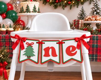 Peut inclure: Une chaise haute blanche ornée d'une bannière festive représentant un sapin de Noël vert et les lettres rouges n et e. La bannière est fixée par des rubans de satin rouge de chaque côté, créant un décor de fête pour un premier anniversaire.