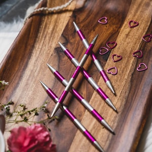 May include: Five pairs of interchangeable knitting needles with purple and silver tips, arranged in a criss-cross pattern on a wooden surface.