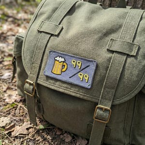 May include: An olive green canvas backpack with adjustable straps and brass buckles. A patch on the front displays a yellow beer mug with white foam and the numbers "99" on a blue background. The backpack is designed for outdoor use.