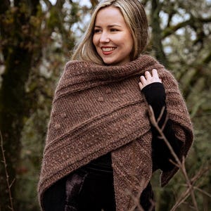 Könnte beinhalten: Ein brauner Strickschal mit strukturiertem Design. Der Schal ist um die Schultern gewickelt. Die Person trägt ein schwarzes Langarmshirt und einen dunklen Rock. Der Hintergrund zeigt kahle Bäume.