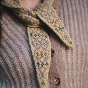 May include: A knitted neckerchief featuring a cream-colored base with a repeating geometric pattern in shades of muted purple, olive green, and brown. The accessory is tied in a simple knot and rests against a textured, light brown knit cardigan.