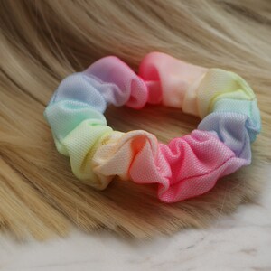 May include: A rainbow-colored scrunchie made of a soft, stretchy fabric. The scrunchie is in a gathered, ruffled style.