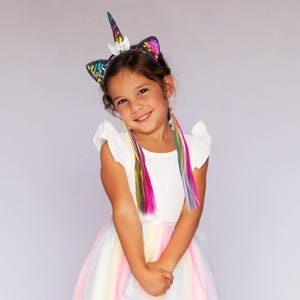 May include: A young girl wearing a rainbow unicorn headband with a sparkly horn and ears. She is wearing a white dress with a rainbow tulle skirt and rainbow hair extensions.