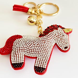 May include: A red and white unicorn keychain with a gold chain and a red tassel. The unicorn is covered in rhinestones.