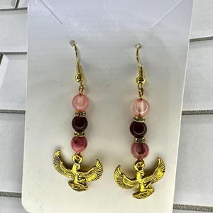 May include: A pair of gold-colored earrings with a dangling design. Each earring features a gold-colored bird pendant, with a series of pink and burgundy beads. The earrings are attached to gold-colored fish hook ear wires.