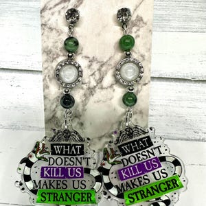 May include: A pair of dangle earrings with a green, white, and black design. The earrings feature a round, white, and silver charm with rhinestones. The bottom of the earrings feature a black and white striped design with the text "What Doesn't Kill Us Makes Us Stranger" in purple and green.