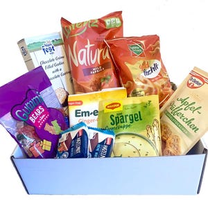 Può includere: Una scatola bianca piena di vari snack confezionati. Gli articoli includono sacchetti di caramelle gommose, patatine alla paprika, biscotti e preparati per zuppe. La confezione presenta testo in più lingue.