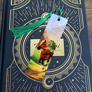 May include: A rectangular bookmark featuring a printed illustration of a character in green clothing riding a brown horse through a landscape, topped with a green silk tassel attached through a circular hole at the top of the bookmark.