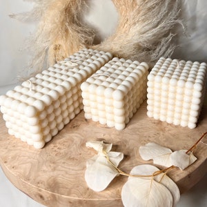 Könnte beinhalten: Drei weiße Kerzen in Säulenform mit einer strukturierten, perligen Oberfläche. Die Kerzen sind in einer Reihe auf einer Holzoberfläche angeordnet.