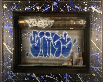 May include: A framed artwork featuring a blue and white graffiti design on a metal garage door. The frame is black with blue and white paint splatters.