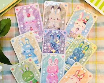 May include: Nine colorful cards with glitter and holographic designs featuring various illustrations of bunnies, a cactus, a mushroom, and a sky. Each card has a title, such as "Cactus Plant", "Sakura Flor", and "Ocean Breeze".