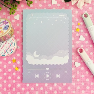 May include: A pastel notepad with a cloud and moon design, featuring a play button and music controls. The notepad has a scalloped edge and a grid pattern. Decorative washi tape and star-shaped embellishments are also visible.