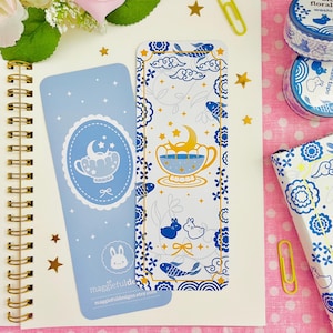 May include: Two blue and white bookmarks with whimsical illustrations of a teacup with a crescent moon, stars, and fish. The bookmarks are decorated with a border of floral and cloud designs. The text "maggiefuldesigns.etsy.com" is visible.