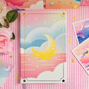 May include: A rectangular notebook with a celestial design. The cover features a yellow crescent moon, stars, and clouds against a pink and blue gradient background. Gold accents frame the design. Decorative star-shaped confetti is scattered around the notebook.