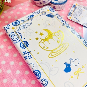 May include: A white notebook featuring a gold crescent moon and star design. The notebook has a blue and gold border with floral and fish motifs. A blue rabbit and bow are also featured on the cover.