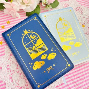 May include: Two notebooks with gold foil accents. One is navy blue, the other is light blue. Both feature a window design with a moon, stars, and clouds. The covers have a scalloped border and a ribbon bookmark.
