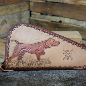 May include: A brown leather case with a detailed image of a dog in a field. The case has a decorative border and a stitched edge. The dog is a reddish-brown color. The case also features crossed rifles and the initials "MC".