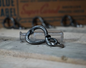 Può includere: Un apribottiglie a forma di cuore argentato con un anello attaccato. L'apribottiglie è appoggiato su una superficie di legno accanto a una bottiglia di vetro trasparente.