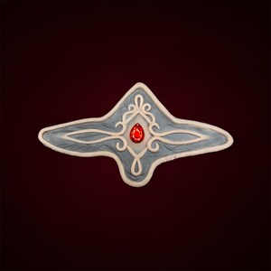 May include: A grey and white ceramic ornament with a red gem in the centre. The ornament is shaped like a star with intricate details.