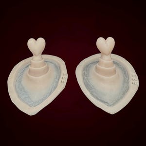 May include: Two matching, off-white, heart-shaped decorative objects. Each has a heart-shaped top and a tiered base, set within a light blue, oval-shaped basin. The background is a dark maroon color.