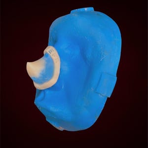 Puede incluir: Una máscara facial de plástico azul con una pieza nasal blanca. La máscara tiene una correa en el lateral.