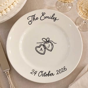 May include: A white ceramic plate featuring black text that reads The Emiles at the top and 24 October 2026 at the bottom. In the center, there is a black line drawing of two connected heart-shaped lockets with the letters A and J inside.