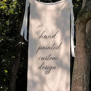 May include: A rectangular off-white fabric banner featuring the black cursive text hand painted custom design. The banner is suspended by white ribbon ties at the top corners, displaying a smooth texture and clean edges against a natural outdoor setting.