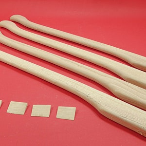 May include: Four unfinished wooden axe handles on a red background. The handles are all the same size and shape, and they are arranged in a row. There are five small wooden squares in front of the handles.