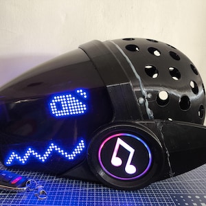 May include: A black, futuristic-looking helmet with a rounded top and a visor-like front. The front displays a digital face with blue LED lights forming eyes and a mouth. A remote control and key ring are in front of the helmet. A musical note is on the side.