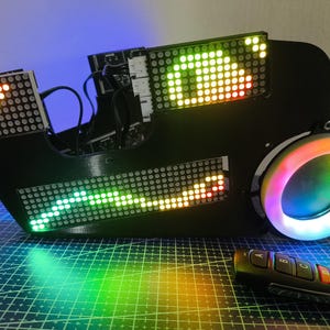 May include: A black electronic device featuring three distinct LED dot matrix display panels showing colorful light patterns, a circular RGB light ring on the right side, and a small black remote control with buttons labeled A, B, C, and D.